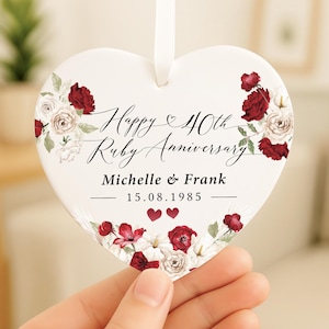 May include: A heart-shaped ceramic ornament with a white background, decorated with red and white floral patterns. The ornament displays the text "Happy 40th Ruby Anniversary", names, and a date.