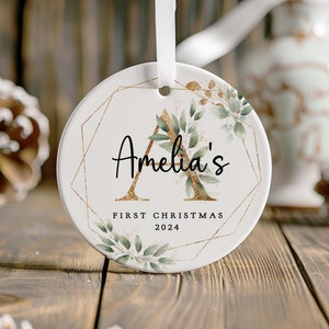 May include: A white ceramic Christmas bauble with a gold geometric design and green foliage. The bauble features the letter "A" with the name "Amelia's" and the text "First Christmas 2024".