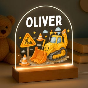 May include: A night light with the name "OLIVER" in white lettering. The light features a cartoon illustration of a yellow excavator, traffic cones, and a construction sign. The base is made of wood.