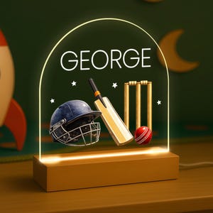May include: A cricket-themed night light, featuring the name "GEORGE". The design includes a cricket helmet, bat, ball, and wickets. The light has a wooden base and a glowing arched top. The light is suitable for a child's bedroom.