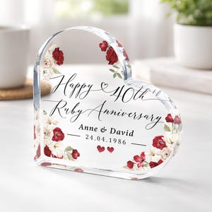 May include: A clear, heart-shaped acrylic ornament with floral detailing. The text reads "Happy 40th Ruby Anniversary" with names and a date. Red and white roses adorn the edges, creating a romantic and celebratory aesthetic.
