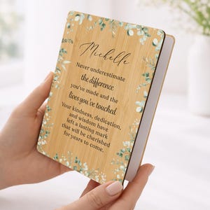 May include: A wooden plaque with the name "Michelle" and a heartfelt message. The plaque has a light brown bamboo-like texture, adorned with delicate green and white floral accents. The text reads, "Never underestimate the difference you've made..."