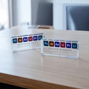 May include: Two clear acrylic desk nameplates with the names Matthew Rowe and Oliver Roberts. Each nameplate has the Adobe Creative Suite icons for Photoshop, Illustrator, After Effects, InDesign, Premiere Pro, and Lightroom. Matthew Rowe's nameplate says "Motion Graphics Designer" and Oliver Roberts' nameplate says "Graphic Designer".