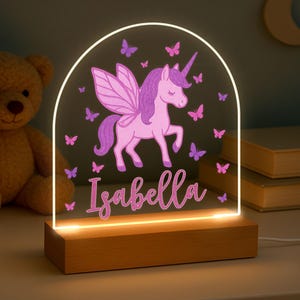 May include: A personalised night light with a pink unicorn, purple wings, and butterflies. The name "Isabella" is written in cursive. The light sits on a wooden base and has a glowing white arch.
