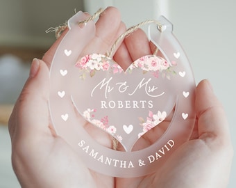 May include: A white horseshoe-shaped acrylic ornament with a pink floral heart design. The ornament features the text "Mr & Mrs Roberts" and "Samantha & David".