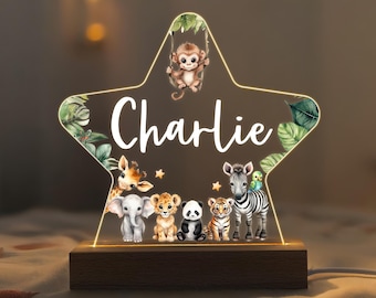 Puede incluir: Una luz nocturna de madera con forma de estrella con un panel de acrílico transparente con un tema de animales de la selva. El panel tiene una inscripción blanca que dice "Charlie" y representa un mono balanceándose en una enredadera, una jirafa, un elefante, un león, un panda, un tigre y una cebra. La luz se ilumina desde abajo.
