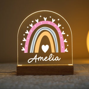 May include: A personalised rainbow night light with a wooden base. The rainbow is in shades of blue, orange, and yellow with pink hearts around it. The name "Amelia" is written below the rainbow.
