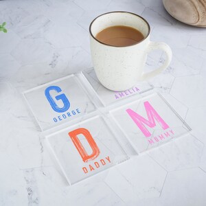 May include: Four clear acrylic coasters with colourful letters and names. The coasters feature the letters G, D, M, and A, with the names George, Daddy, Mummy, and Amelia printed below each letter.