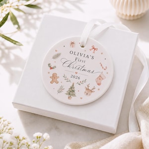 May include: A round white ceramic ornament featuring the text Olivia's First Christmas 2026. The design includes watercolour illustrations of a gingerbread man, a reindeer, a Christmas tree, a stocking, a gift, a bow, and holly leaves.