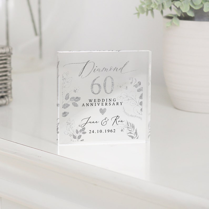 K&ouml;nnte beinhalten: Ein klarer Glasblock mit einem silbernen Glitzerdesign. Der Text "Diamond 60 Wedding Anniversary June & Ron 24.10.1962" ist auf dem Block gedruckt.