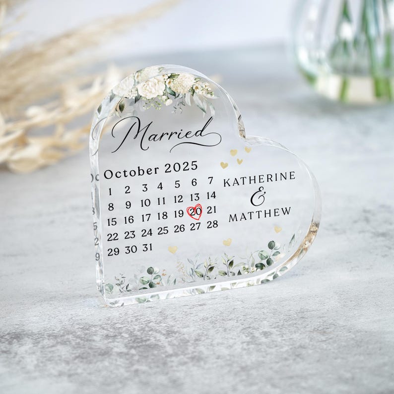 May include: A clear heart-shaped glass paperweight with a floral design and the words "Married" and "October 2025" printed on it. The date "20" is highlighted in red. The names "Katherine & Matthew" are printed below the calendar.