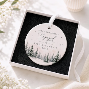 May include: A round white ceramic ornament with a white ribbon, presented in a black-lined gift box. The ornament features a watercolour forest scene and black text reading FIRST CHRISTMAS Engaged, MILLIE & ARCHIE, and the year 2026.