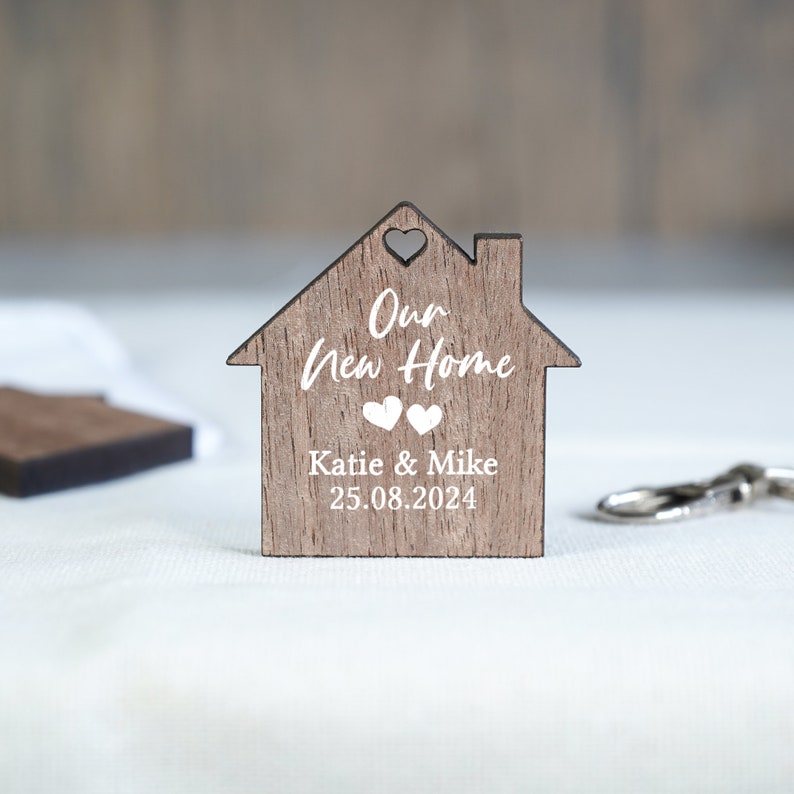 May include: A wooden house-shaped keychain with the text "Our New Home" and the names "Katie & Mike" and the date "25.08.2024".