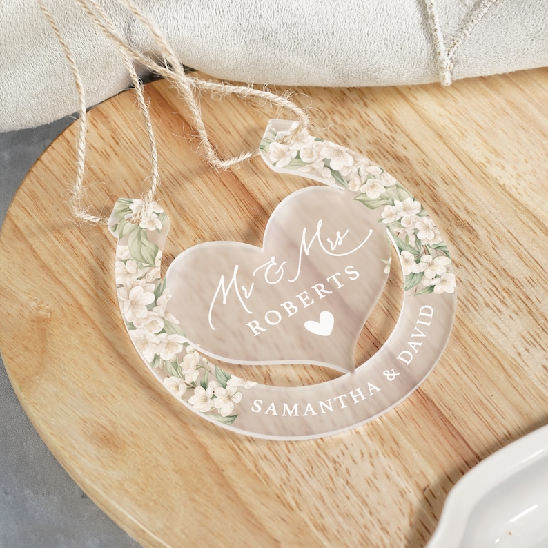 May include: A clear acrylic horseshoe with a floral design and a heart-shaped cutout. The heart cutout has the text "Mr & Mrs Roberts" and a small heart. The horseshoe has the text "Samantha & David" on the bottom.