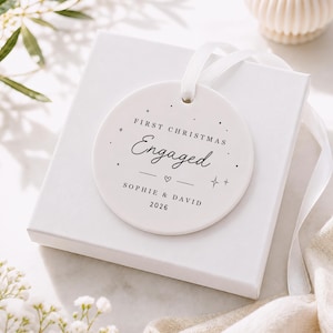 May include: A round white ceramic ornament with a white ribbon, featuring the black printed text FIRST CHRISTMAS Engaged, the names SOPHIE & DAVID, and the year 2026, decorated with small star and heart symbols, resting on a white gift box.