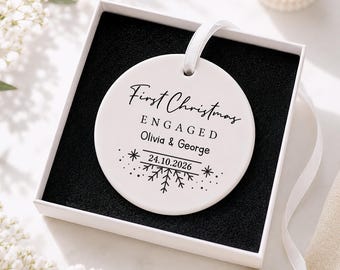 May include: A round white ceramic ornament featuring black text that reads First Christmas ENGAGED Olivia & George 24.10.2026 with a snowflake graphic. The ornament hangs by a white ribbon inside a square white gift box with black foam lining.