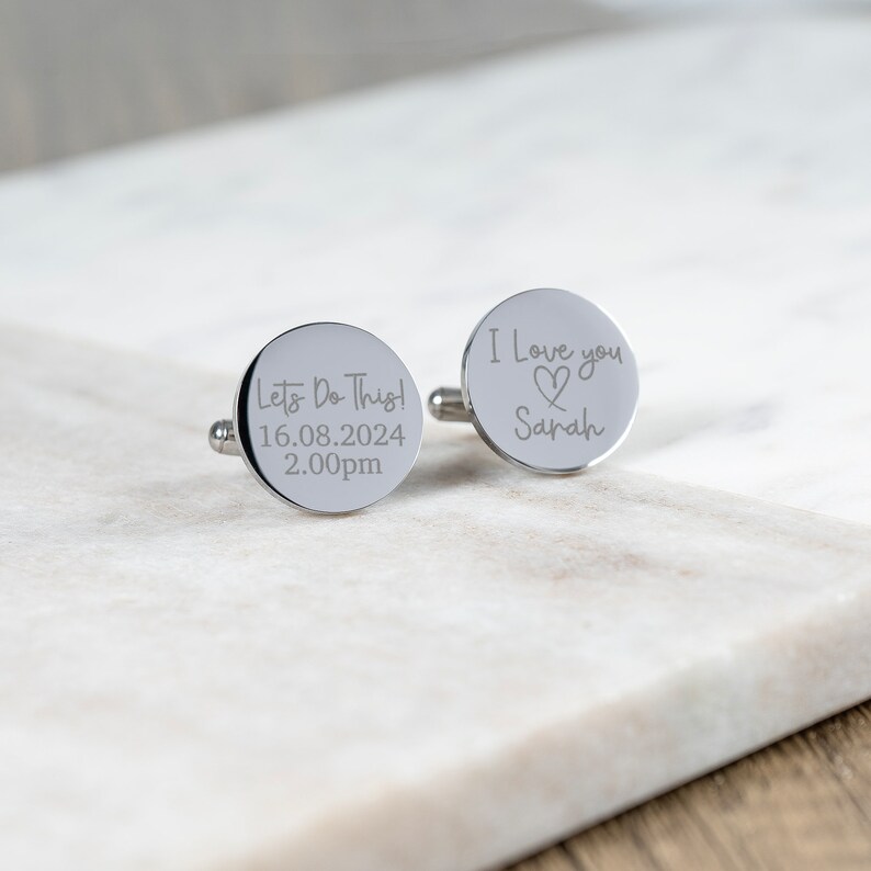 May include: Two silver cufflinks with engraved text. One cufflink says "Let's Do This! 16.08.2024 2.00pm". The other cufflink says "I love you" with a heart symbol and "Sarah".