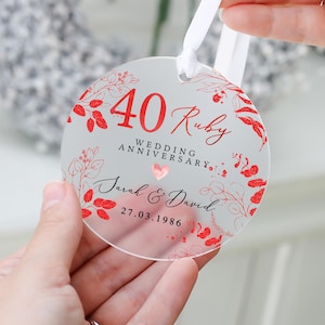 May include: A clear, round ornament with red text and floral accents. The ornament commemorates a 40th Ruby Wedding Anniversary, with the names "Sarah & David" and the date "27.03.1986" displayed. A white ribbon is attached for hanging.