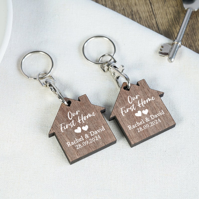 May include: Two wooden house-shaped keychains with silver key rings. The keychains are engraved with the text "Our First Home" and the names "Rachel & David" and the date "28.09.2024".