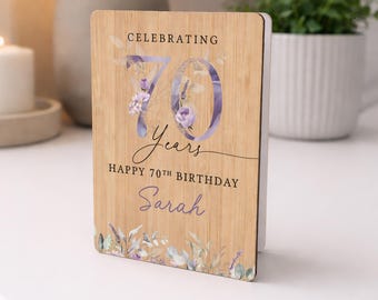May include: A wooden birthday card with the text "Celebrating 70 Years Happy 70th Birthday Sarah". The number 70 is in a purple floral design, with additional floral accents at the top and bottom. The card is standing upright.