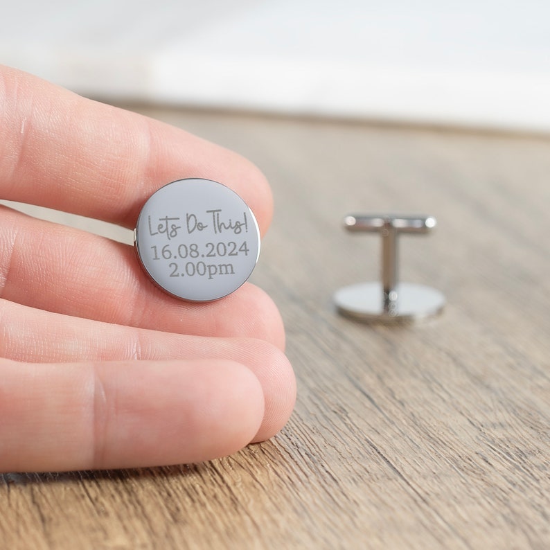 May include: Silver cufflinks with a round, engraved design. The engraving reads "Let's Do This! 16.08.2024 2.00pm".