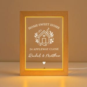 May include: A rectangular wooden framed LED light with the words "HOME SWEET HOME" and a house graphic. Below the graphic is the text "24 APPLEWAY CLOSE" and the names "Rachel & Matthew". A white heart is at the bottom.