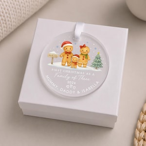 May include: A round, frosted acrylic Christmas ornament featuring a printed illustration of three gingerbread figures, a North Pole sign, and a Christmas tree. The text reads: FIRST CHRISTMAS AS A FAMILY OF THREE, 2026, MUMMY, DADDY & ISABELLA.