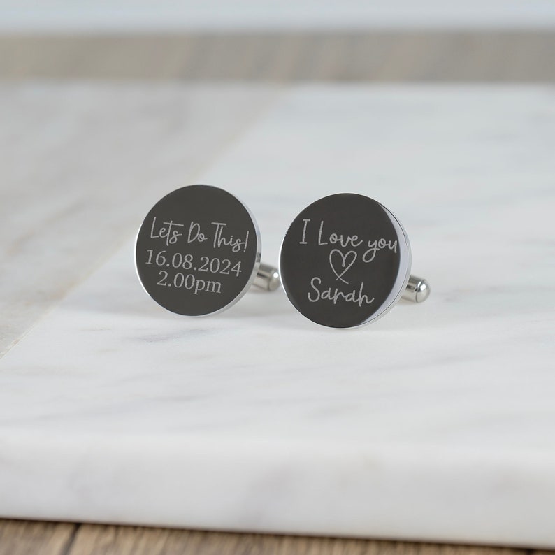 May include: A pair of silver cufflinks with engraved messages. One cufflink says "Let's Do This! 16.08.2024 2.00pm" and the other says "I Love You" with a heart and the name "Sarah".