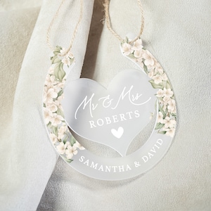 May include: White horseshoe-shaped wedding decoration with a heart-shaped cutout in the center. The heart has the text "Mr & Mrs Roberts" and a small heart at the bottom. The horseshoe is decorated with white flowers and green leaves. The bottom of the horseshoe has the text "Samantha & David".