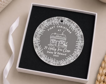 May include: A circular clear acrylic ornament featuring a white etched design of a house, a leafy wreath border, and the text First Christmas at 29 Holly Tree Close, Daisy & Edward, 2026. The ornament is presented inside a square white gift box.