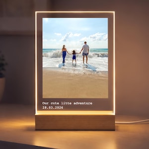 May include: A rectangular LED light with a family photo on the front. The photo shows a family of three at the beach. The light has a wooden base and the text "Our cute little adventure 28.03.2026" is displayed.