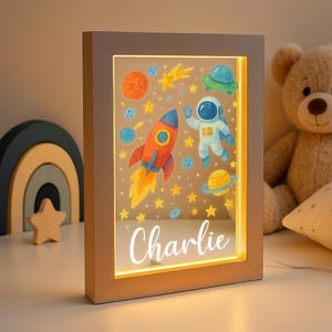 May include: A rectangular, illuminated night light with a space theme. The artwork features a rocket, astronaut, planets, and stars. The name "Charlie" is written in cursive at the bottom. The light has a wooden frame and is plugged in.