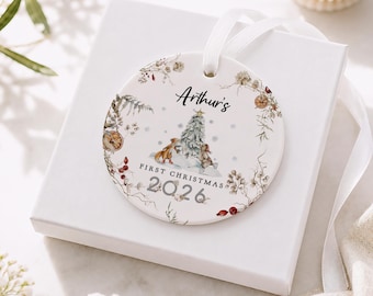 May include: A round white ceramic Christmas ornament featuring a watercolor illustration of a fox and a rabbit near a tree. The text reads Arthur's FIRST CHRISTMAS 2026. The design includes dried orange slices, berries, and foliage, with a white ribbon.