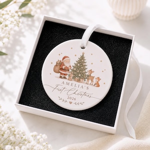 May include: A round white ceramic Christmas ornament featuring a printed illustration of Santa Claus, a decorated pine tree, a rabbit, a deer, and a fox. The text reads AMELIA'S First Christmas 2026. It includes a white ribbon and a black gift box.