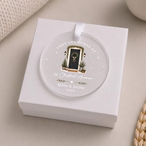 May include: A circular frosted acrylic ornament with a white ribbon, featuring a printed illustration of a black front door with a wreath, flanked by small trees. Text reads: FIRST CHRISTMAS AT 26 Chestnut Avenue Millie & Archie 2026.