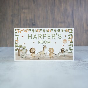 May include: A white wooden sign with a green leafy border and a peach-coloured moon. The sign says "Harper's Room" and features a line of cartoon animals including an elephant, lion, giraffe, tiger, zebra, and monkey.