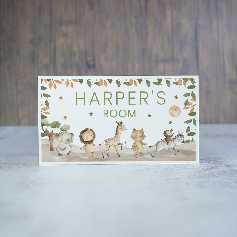 May include: A white wooden sign with a green leafy border and a peach-coloured moon. The sign says "Harper's Room" and features a line of cartoon animals including an elephant, lion, giraffe, tiger, zebra, and monkey.