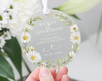 May include: A round, clear glass ornament with a white daisy floral design. The ornament has the text "Wendy & Jonathon Wedding Anniversary 10.07.1992" printed on it.