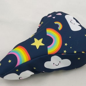 Könnte beinhalten: Ein blauer Fahrradsattelbezug mit einem Regenbogen-, Wolken- und Sternenmuster. Der Bezug hat einen Riemen mit einem Metallring.