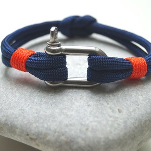 Könnte beinhalten: Ein blaues und orangefarbenes Seil-Armband mit einem silbernen Metallverschluss. Das Armband ist mit einem Knoten gebunden und hat einen kontrastierenden orangefarbenen Streifen.