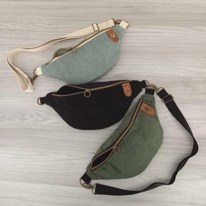 Könnte beinhalten: Drei Gürteltaschen aus Kord mit verstellbaren Riemen und Reißverschlüssen. Eine ist hell salbeigrün mit cremefarbenem Riemen, eine schwarz mit braunem Lederetikett mit der Aufschrift J.S. und eine olivgrün mit schwarzem Riemen und Blumenemblem.