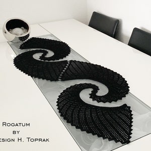 Puede incluir: Un camino de mesa de crochet negro con un diseño en espiral. El camino de mesa está sobre una mesa blanca con dos sillas negras en el fondo. El texto "Rogatum by HT Design H. Toprak" está impreso en la mesa.