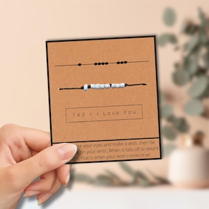 May include: A brown card with two black string bracelets. The top bracelet has a series of black dots, and the bottom bracelet has light blue beads. The card reads "143 = I Love You".