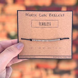 May include: A black string bracelet with silver beads spelling out the word "FEARLESS" in Morse code. The bracelet is displayed on a brown card with the words "Morse Code Bracelet" and "FEARLESS" printed on it. The card also includes the website "icametoworship.com" and the phrase "Even in the Valley Gifts".