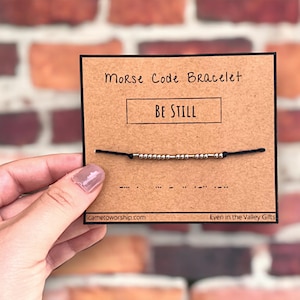 May include: A brown card with the text "Morse Code Bracelet" and "BE STILL" in black ink. A black string bracelet with silver beads is displayed on the card. The Morse code for "BE STILL" is shown below the bracelet.