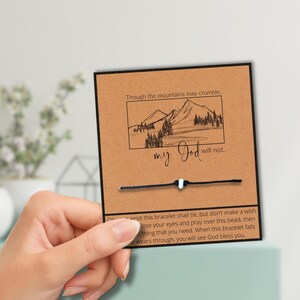 Puede incluir: Una pulsera de cuerda negra con una cuenta blanca se muestra en una tarjeta marrón. La tarjeta tiene una ilustración de montaña y el texto "Though the mountains may crumble, my God will not."