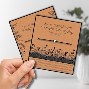 May include: Two cards with a black string bracelet. The cards are brown with black text that reads "She is clothed with strength and dignity. Proverbs 31:25." The cards have a floral design and a bracelet with a light blue bead.