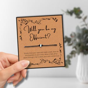 May include: A brown card with the text "Will you be my Officiant?" in elegant script. The card features a black string bracelet with a white bead. Decorative floral elements frame the text. The card is held by a hand.