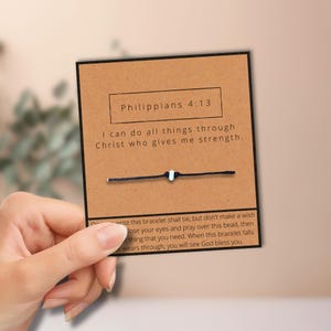 May include: A card with the text "Philippians 4:13" and the message "I can do all things through Christ who gives me strength." A black string bracelet with a white bead is attached to the card. The card is held by a hand.