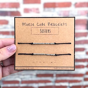 Op de afbeelding: Twee zwarte armbandjes van touw met zilveren kralen die het woord "SISTERS" in morsecode spellen. De armbandjes worden getoond op een bruine kaart met de tekst "Morse Code Bracelets" en "Even in the Valley Gifts".